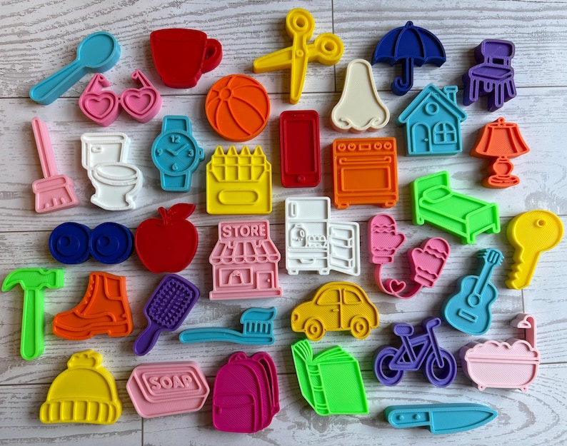 May include: A collection of colorful plastic cookie cutters in various shapes including a spoon, cup, scissors, umbrella, chair, glasses, ball, nose, house, lamp, broom, toilet, watch, crayons, phone, oven, bed, apple, store, fridge, mittens, key, hammer, boot, brush, toothbrush, car, guitar, hat, soap, backpack, book, bicycle, bathtub, and knife.