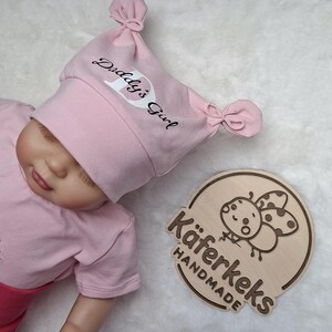 Könnte beinhalten: Eine hellrosa Babymütze mit zwei geknoteten oberen Ecken und dem in Schwarz und Weiß gedruckten Text Daddy's Girl. Daneben liegt eine runde Holzplakette mit einer Marienkäfer-Illustration und dem eingravierten Text Käferkeks HANDMADE.