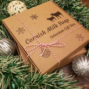 May include: A brown cardboard gift box tied with red and white twine, labeled "Cornish Milk Soap Selection Gift Box." The box is decorated with snowflake and cow illustrations. Christmas ornaments and green garland surround the box.