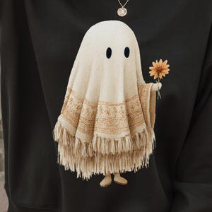 May include: Black sweatshirt featuring a whimsical ghost design. The ghost is draped in a cream-colored shawl with a fringed hem and holds a yellow sunflower. The design is centered on the front of the sweatshirt.