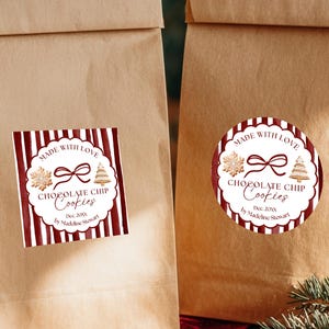May include: Two brown paper bags with festive labels that read "Made with Love Chocolate Chip Cookies." The labels feature a red and white striped design with a bow and Christmas tree illustration. The bags are on a wooden surface.
