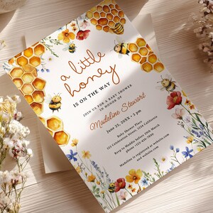 May include: A baby shower invitation with a watercolour design. The invitation features the text "a little honey is on the way" with illustrations of bees, honeycombs, and flowers. The invitation includes the name Madeline Stewart and event details.