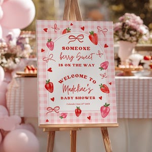 Puede incluir: Un letrero a cuadros rosa y blanco sobre un caballete de madera. El letrero dice "Someone berry Sweet is on the way. Welcome to Madeline's Baby Shower." Está decorado con fresas rojas, lazos y corazones. El evento es en Colorado.