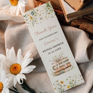 May include: A bookmark with the text "Thank You for Celebrating Emma's Bridal Shower 06.25.20XX". The bookmark features a floral design with daisies and a stack of books tied with a pink ribbon. Daisies and old books are in the background.