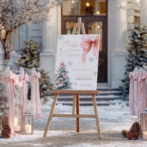 Op de afbeelding: Een houten ezel toont een babyborrelbord met een winterthema. Het bord heeft een roze strik, sneeuwvlokken en een illustratie van een kerstboom. De tekst op het bord luidt "Baby It's Cold Outside" en "Welcome to Madeline's Baby Shower."