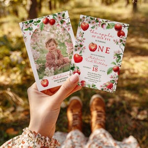 May include: Two invitations for a first birthday celebration. The invitations feature a photo of a baby surrounded by flowers and apples. Text includes "Madeline's First Birthday" and "the apple of our eye is turning ONE."