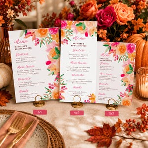 May include: Three white menu cards for Madeline's Bridal Shower, displayed in gold circular stands. Each card features watercolor floral illustrations in orange, pink, and green, with text listing starters, main courses, desserts, and drinks.