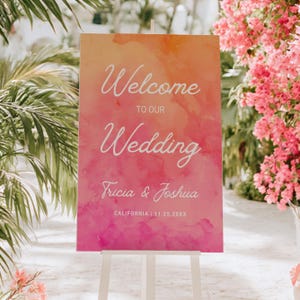 May include: A rectangular wedding welcome sign featuring a watercolor gradient background transitioning from orange at the top to pink at the bottom. The white text reads Welcome to our Wedding, Tricia & Joshua, and CALIFORNIA | 11.25.20XX.