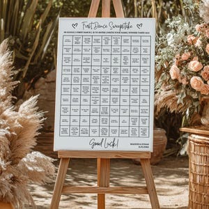 May include: A white canvas sign on a wooden easel, featuring a "First Dance Sweepstake" game. The sign includes a grid of song titles, instructions, and the text "Good Luck!". The names "Maddie & Ethan" and a date are also present.