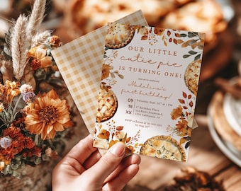 May include: A birthday invitation with the text "OUR LITTLE cutie pie IS TURNING ONE!" features pie illustrations and floral accents. The invitation is held with a gingham envelope, and the background includes a floral arrangement and a pie.