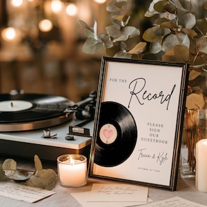 Könnte beinhalten: Ein gerahmtes Schild mit dem Text „For the Record“ und „Please Sign Our Guestbook“. Eine Vinyl-Schallplatten-Grafik befindet sich auf dem Schild. Eine brennende Kerze und ein Plattenspieler sind im Hintergrund.