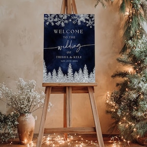以下が含まれることがあります： 木製のイーゼルには、ネイビーブルーのウェディングウェルカムサインが表示されています。サインには、銀色の雪の結晶と森のデザインが施されています。サインのテキストは「WELCOME TO THE wedding OF TRISHA & KYLE NOV. 25TH 20XX」です。