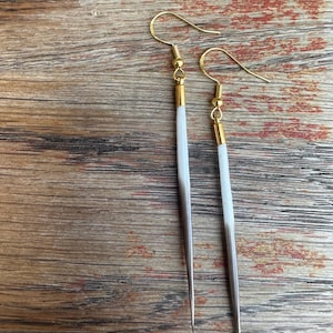 May include: A pair of handmade earrings with a minimalist design. Each earring features a gold-colored hook and a slender, white and silver-toned pendant. The earrings are displayed against a wooden background.