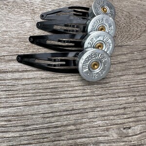 Może przedstawiać: Cztery czarne metalowe spinki do włosów z srebrnymi łuskami po nabojach Winchester 72 GA. Akcesoria do włosów ułożone są na drewnianej powierzchni.