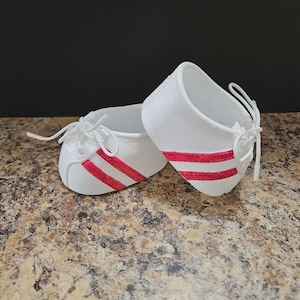 May include: A pair of white baby shoes with red stripes and white laces. The shoes are made of a soft material and are designed for infants. The shoes are placed on a brown and tan speckled surface.
