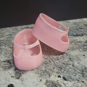 May include: A pair of light pink doll shoes featuring a rounded toe, a single strap across the top, and a low-cut heel design. The shoes are molded from a smooth, solid material and display a subtle textured pattern on the front toe area.