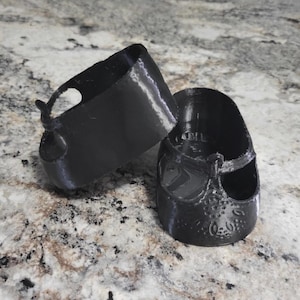 May include: Two black 3D-printed baby shoes. One shoe is upright, and the other is on its side. The shoes have a Mary Jane style with a strap and buckle. The shoes are sitting on a speckled countertop.