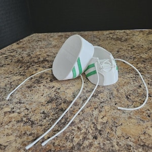May include: A pair of white miniature sneakers with green stripes and white laces. The shoes are made of a smooth material and are designed for a small doll or toy. The sneakers are displayed on a speckled brown surface.