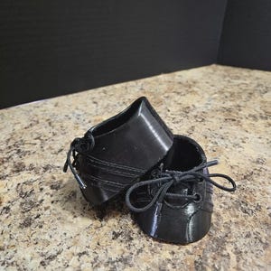May include: Two small, black, 3D-printed shoes with laces. The shoes are designed with a classic style, featuring a rounded toe and a lace-up closure. The shoes are likely for a doll or other small figure.