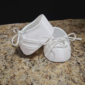May include: A pair of small, white baby shoes with laces. The shoes are made of a smooth material and have a classic sneaker design. The shoes are resting on a speckled surface.