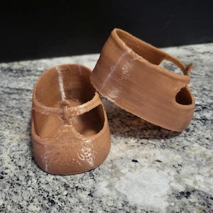 Könnte beinhalten: Ein Paar hellbraune Puppenschuhe mit T-Riemen-Design und einem strukturierten Blumenmuster auf dem abgerundeten Zehenbereich. Die Schuhe haben eine glatte Oberfläche und eine tief ausgeschnittene Öffnung, wobei ein Schuh aufrecht steht.