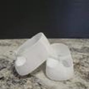 May include: A pair of white baby shoes. The shoes are made of a soft material and have a rounded toe and a strap across the top. The shoes are designed for infants and toddlers.