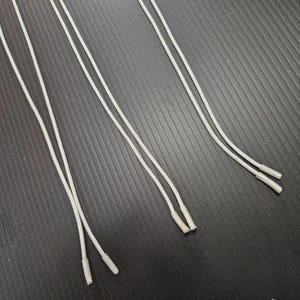 May include: Several pairs of white shoelaces with plastic aglets are arranged diagonally against a dark, textured background. The laces are thin and appear to be new.