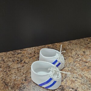 May include: A pair of white baby shoes with blue stripes and white laces. The miniature sneakers are designed to resemble adult athletic shoes. The shoes are displayed on a speckled brown surface with a dark background.