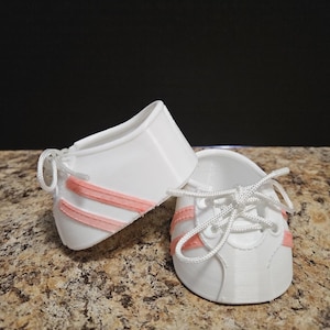 May include: A pair of white miniature sneakers with pink stripes and white laces. The small shoes are designed with a classic athletic style, perfect for a baby or doll. The shoes are displayed on a textured, light brown surface.