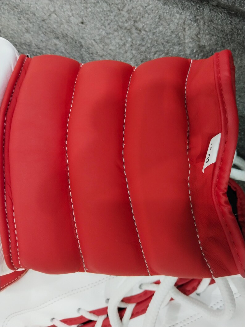 Pu&ograve; includere: Primo piano di una scarpa sportiva rossa e bianca. La scarpa presenta un colletto rosso imbottito con cuciture bianche e una base bianca. I lacci bianchi sono visibili nella parte inferiore dell'immagine.