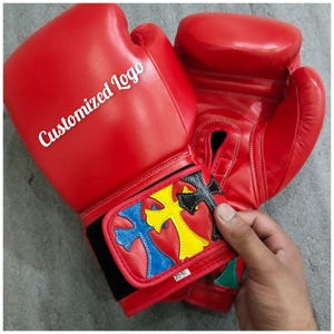 Puede incluir: Un par de guantes de boxeo rojos con el texto "Customized Logo" en el guante izquierdo. La correa de la muñeca presenta un diseño de cruz en azul, amarillo y negro. Los guantes son de 16 oz, aproximadamente 454 gramos.