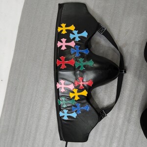 May include: Black leather accessory with colorful cross appliques. The accessory has a curved shape with a strap and buckle. The crosses are in various colors, including yellow, pink, blue, red, and green.