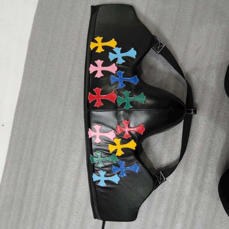 May include: Black leather accessory with colorful cross appliques. The accessory has a curved shape with a strap and buckle. The crosses are in various colors, including yellow, pink, blue, red, and green.