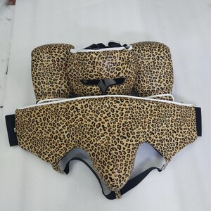 Puede incluir: Un conjunto de boxeo con estampado de leopardo. El conjunto incluye un protector de cabeza, guantes de boxeo y un protector corporal. El estampado de leopardo presenta manchas marrones y beige sobre un fondo beige claro. El conjunto tiene ribetes blancos y correas negras.