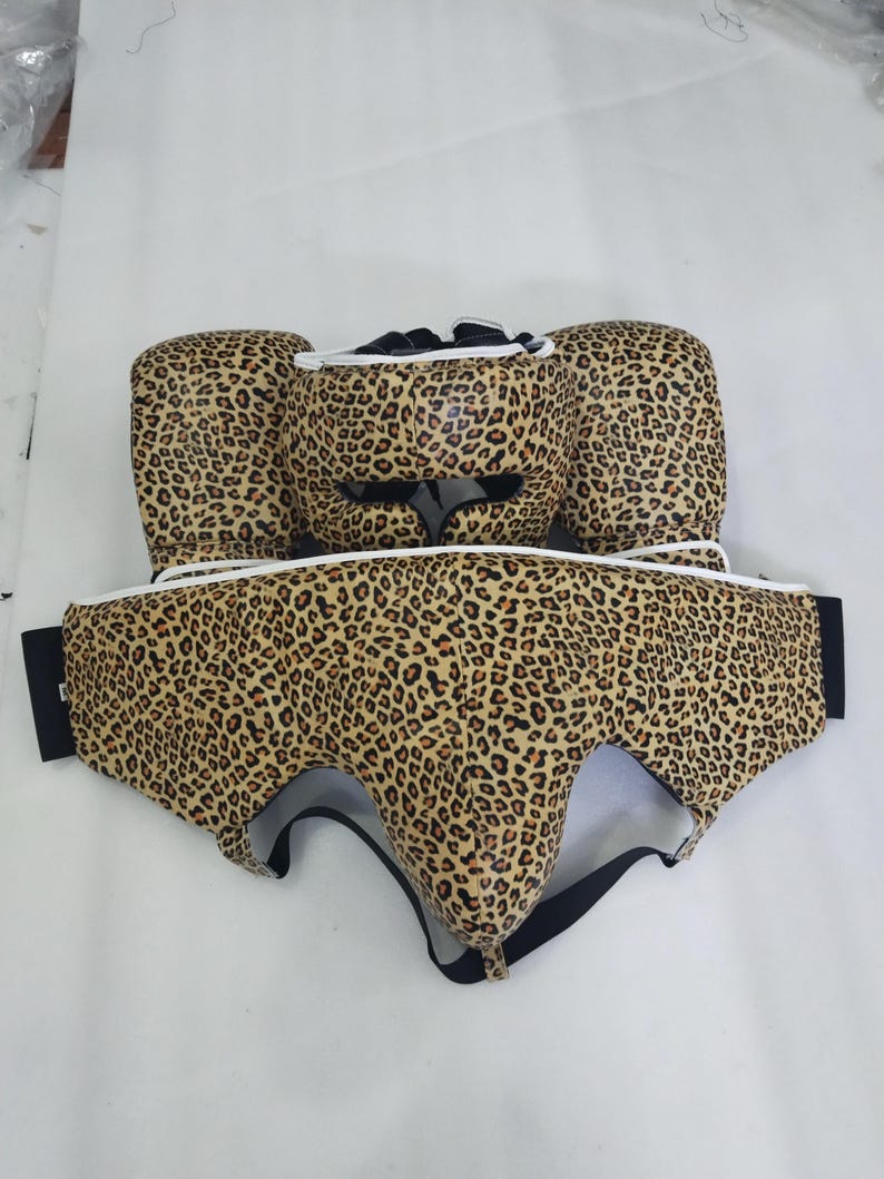 Puede incluir: Un conjunto de boxeo con estampado de leopardo. El conjunto incluye un protector de cabeza, guantes de boxeo y un protector corporal. El estampado de leopardo presenta manchas marrones y beige sobre un fondo beige claro. El conjunto tiene ribetes blancos y correas negras.