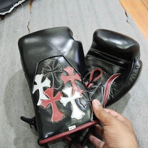 Puede incluir: Guantes de boxeo de cuero negro con cruces decorativas rojas y blancas. Los guantes tienen un diseño con cordones y se muestran sobre una superficie gris. Adecuados para boxeo u otros deportes de combate.