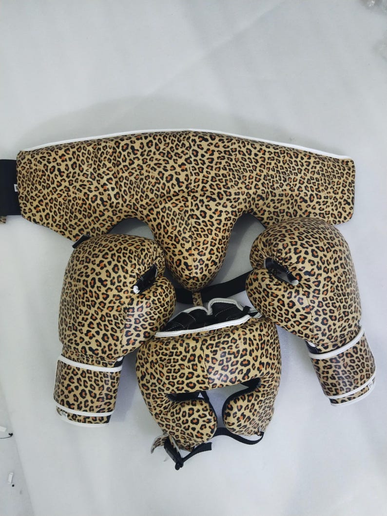 Puede incluir: Conjunto de equipamiento de boxeo con estampado de leopardo. El conjunto incluye guantes de boxeo, protector de cabeza y protector corporal. Los guantes y el casco tienen ribetes blancos. El protector corporal tiene una correa negra.