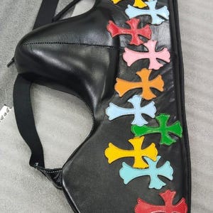 May include: Black leather bag with colorful cross appliques in red, pink, orange, yellow, green, and light blue. The bag has a strap and the letter "M" is visible.