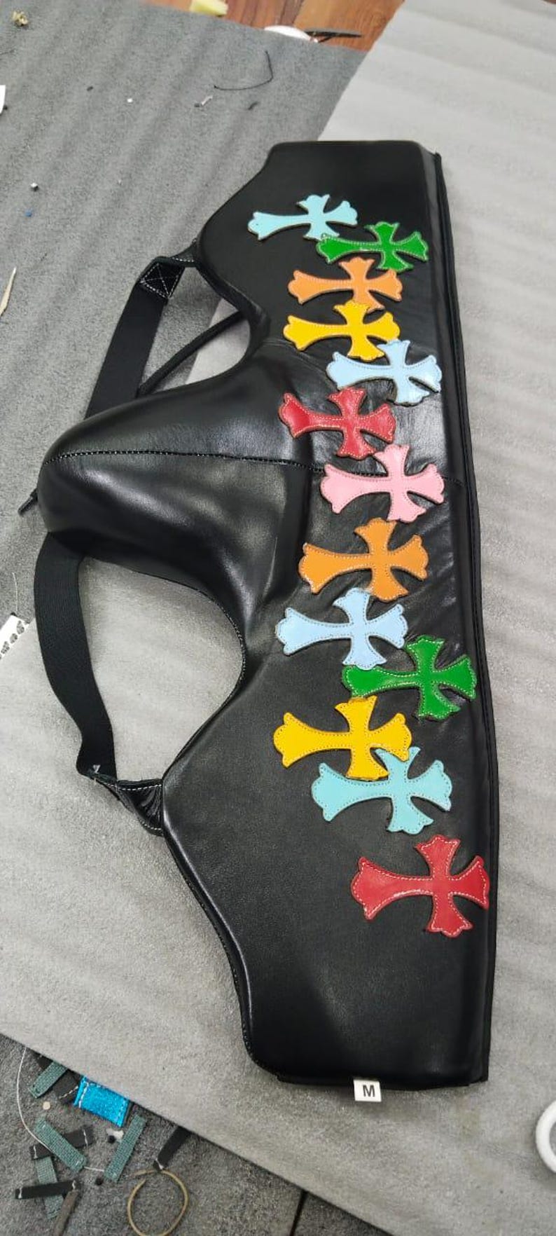 May include: Black leather bag with colorful cross appliques in red, pink, orange, yellow, green, and light blue. The bag has a strap and the letter "M" is visible.