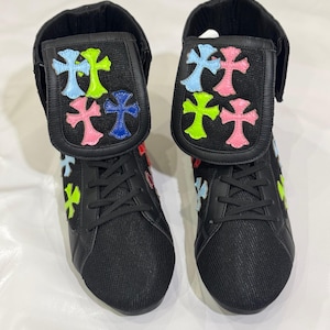 Può includere: Un paio di sneakers alte nere con disegni a croce colorati. Le sneakers presentano un tessuto a rete sulla punta e sui lati, con una patta ripiegabile ornata da croci rosa, blu e verdi. I lacci sono neri.