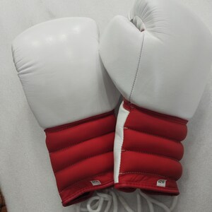 Pu&ograve; includere: Un paio di guantoni da boxe bianchi e rossi. I guanti sono bianchi sulla parte superiore e rossi sulla zona del polso. La zona del polso ha un design trapuntato. Sono visibili lacci bianchi.