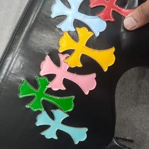 May include: Black leather item adorned with colorful cross-shaped appliques in shades of pink, yellow, green, blue, and red. The crosses are stitched with white thread.