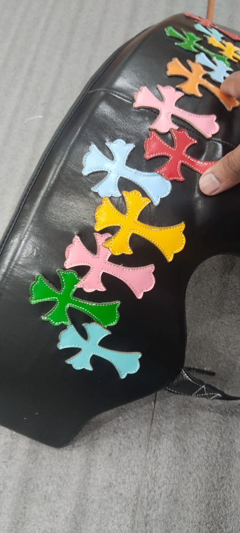 May include: Black leather item adorned with colorful cross-shaped appliques in shades of pink, yellow, green, blue, and red. The crosses are stitched with white thread.