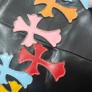 May include: Black leather bag adorned with colorful cross-shaped appliqu&eacute;s. The crosses are in shades of pink, red, orange, yellow, green, and blue, with white stitching. The bag's design is eye-catching and unique.