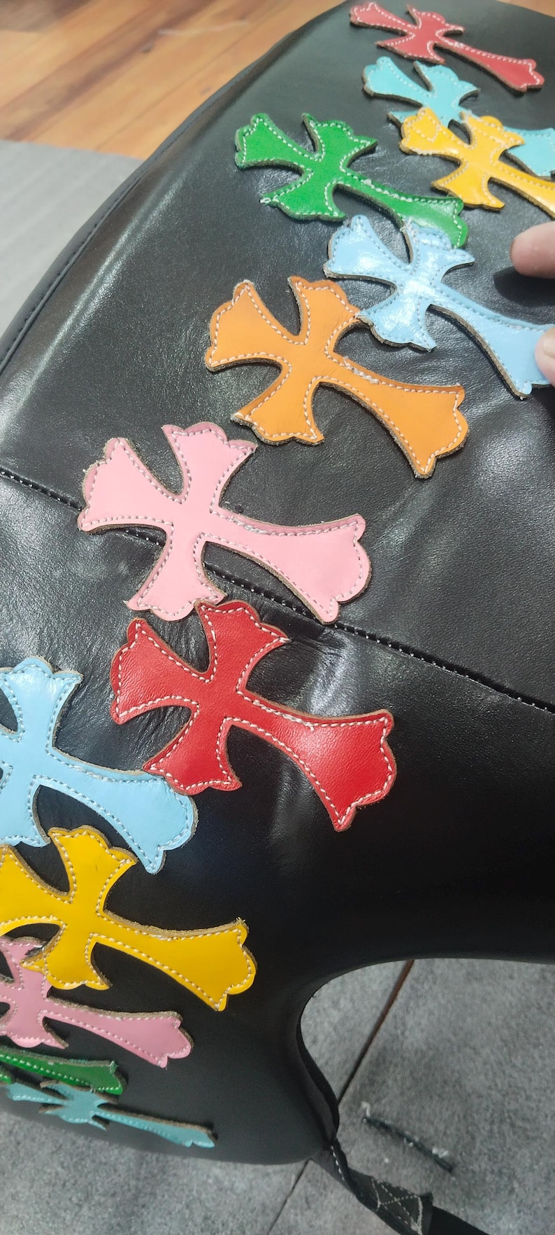 May include: Black leather bag adorned with colorful cross-shaped appliqu&eacute;s. The crosses are in shades of pink, red, orange, yellow, green, and blue, with white stitching. The bag's design is eye-catching and unique.