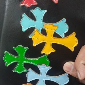May include: A collection of colorful leather cross appliques in red, blue, yellow, green, and orange, stitched onto a black surface. The crosses have a decorative design and are arranged in a pattern.