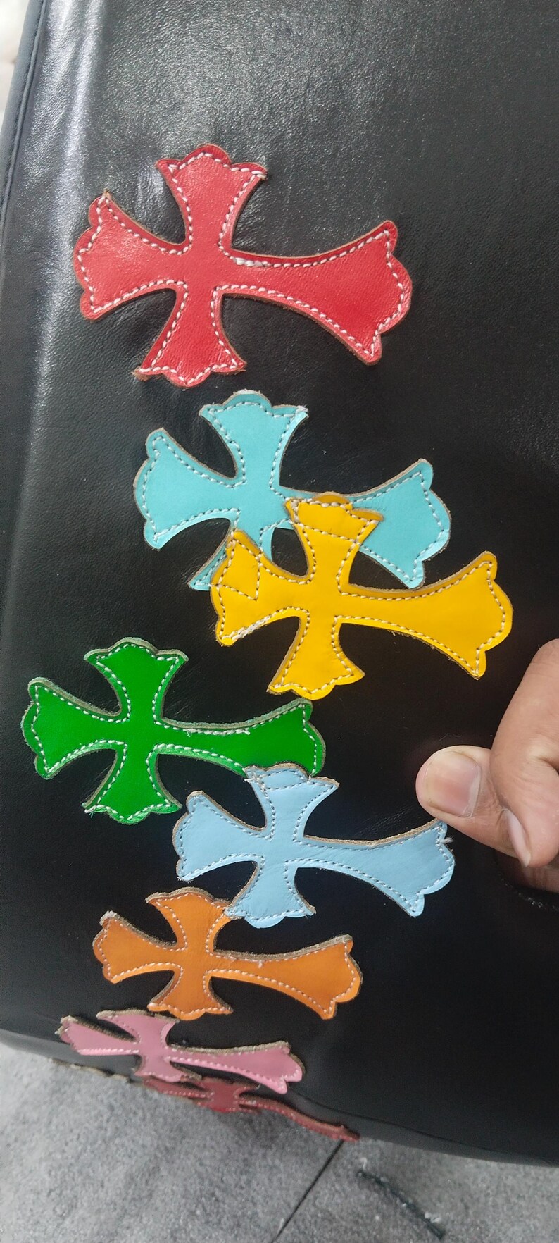May include: A collection of colorful leather cross appliques in red, blue, yellow, green, and orange, stitched onto a black surface. The crosses have a decorative design and are arranged in a pattern.