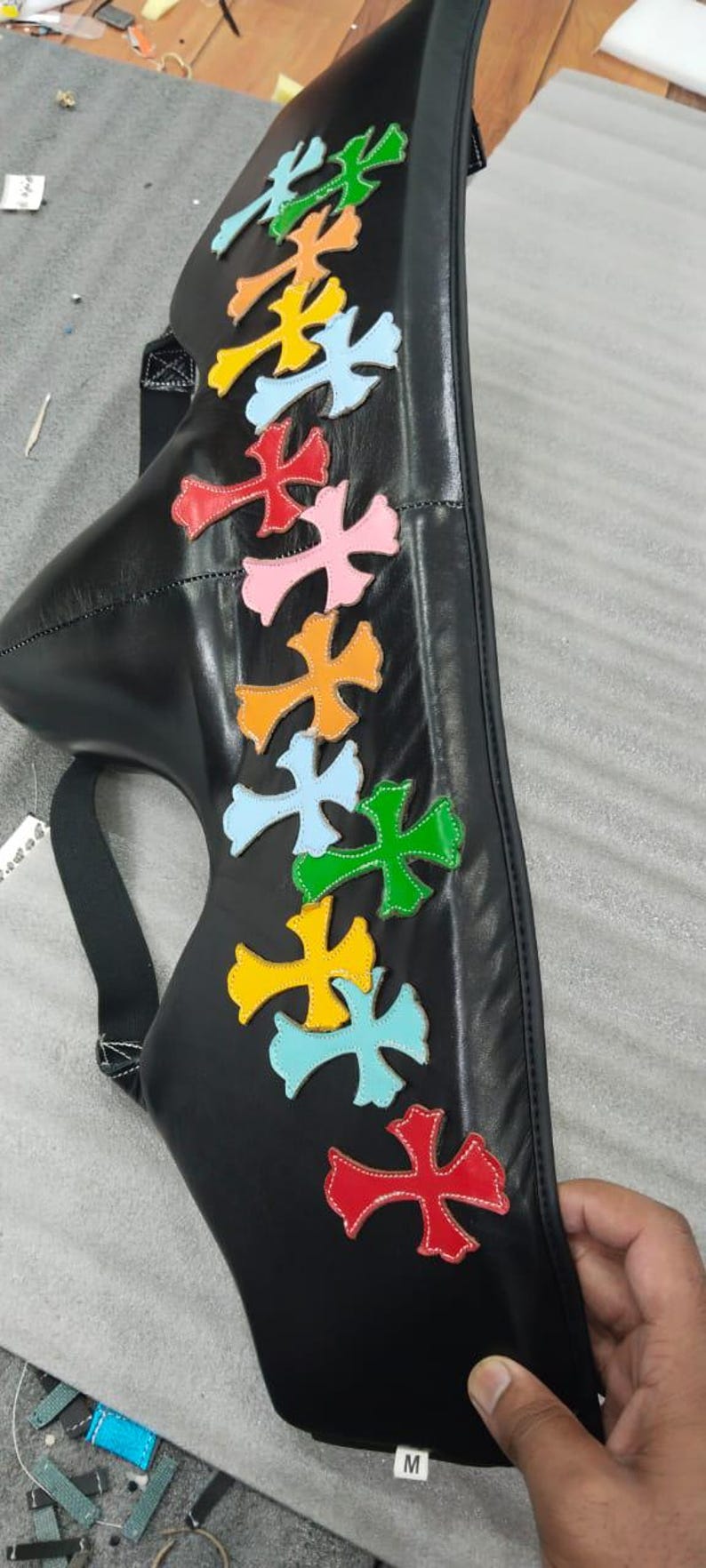 May include: Black leather bag with colorful cross-shaped appliqu&eacute;s in red, pink, orange, light blue, yellow, and green. The bag has a strap and a curved shape, with the letter "M" visible.