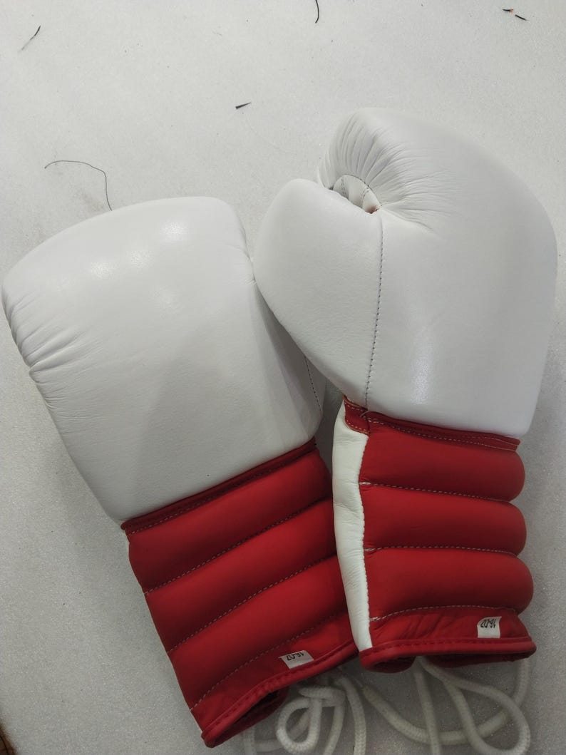 Pu&ograve; includere: Un paio di guantoni da boxe bianchi e rossi. I guantoni hanno una parte superiore imbottita bianca e una zona del polso rossa con un design trapuntato. I lacci bianchi sono visibili in basso.