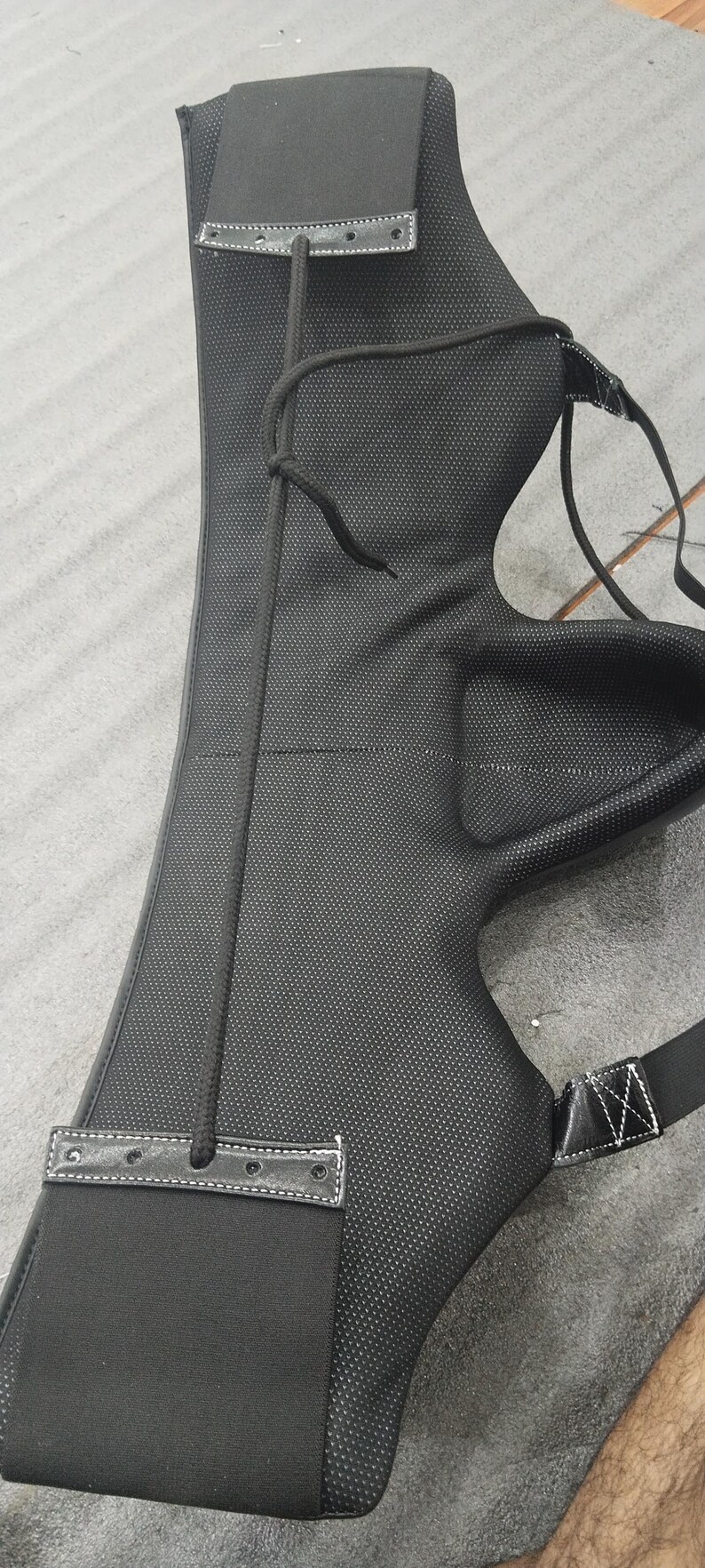 May include: Black archery arm guard with a textured surface and a drawstring. The guard has a padded section and a strap with a white stitched design. The arm guard is designed to protect the forearm.